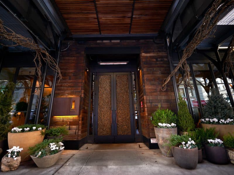 Entrance of 1 Hotel Central Park featuring eco-friendly design, wooden facade, decorative plants, and elegant double doors with intricate patterns.