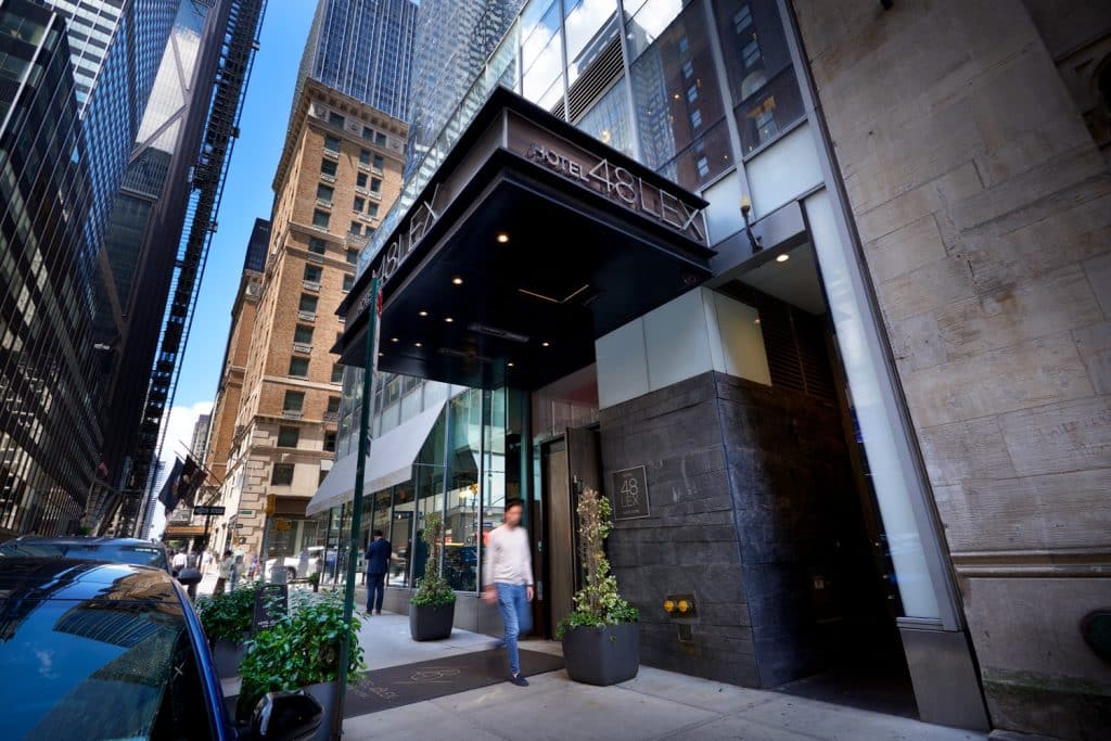 Hotel 48LEX New York entrance with modern architecture, surrounded by Midtown Manhattan buildings, featuring a guest walking in and planters at the entrance.