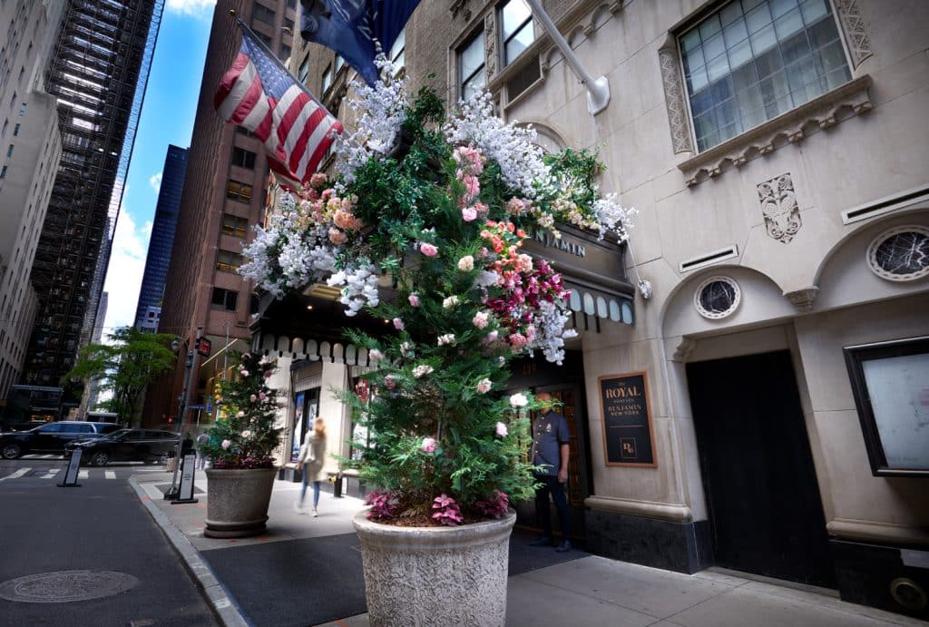 Floral display at The Benjamin Royal Sonesta New York entrance, featuring vibrant flowers and an American flag, with Midtown Manhattan buildings in the background.