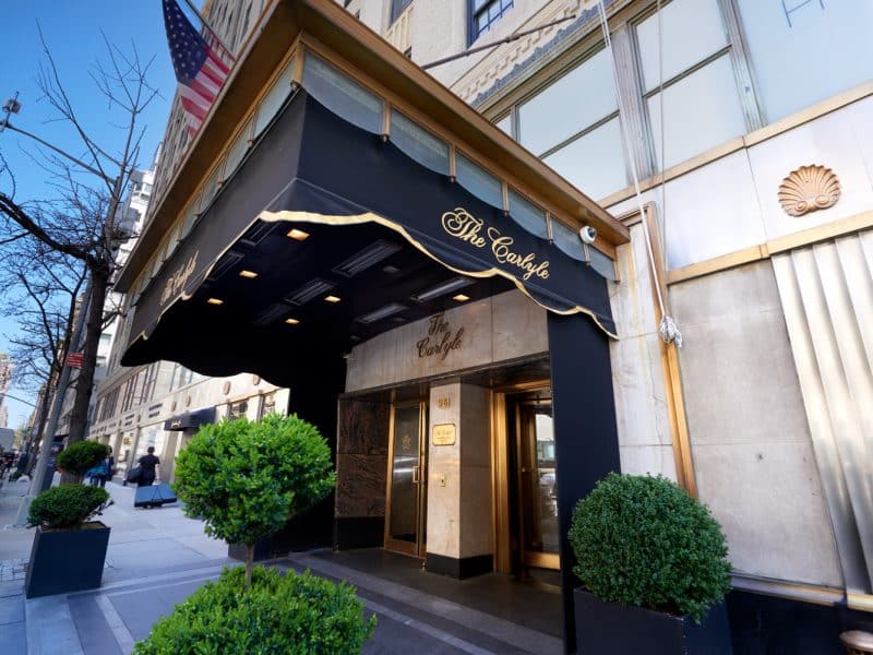 Entrance of The Carlyle, A Rosewood Hotel, featuring elegant black awning with gold lettering, manicured greenery, and upscale Manhattan street setting.