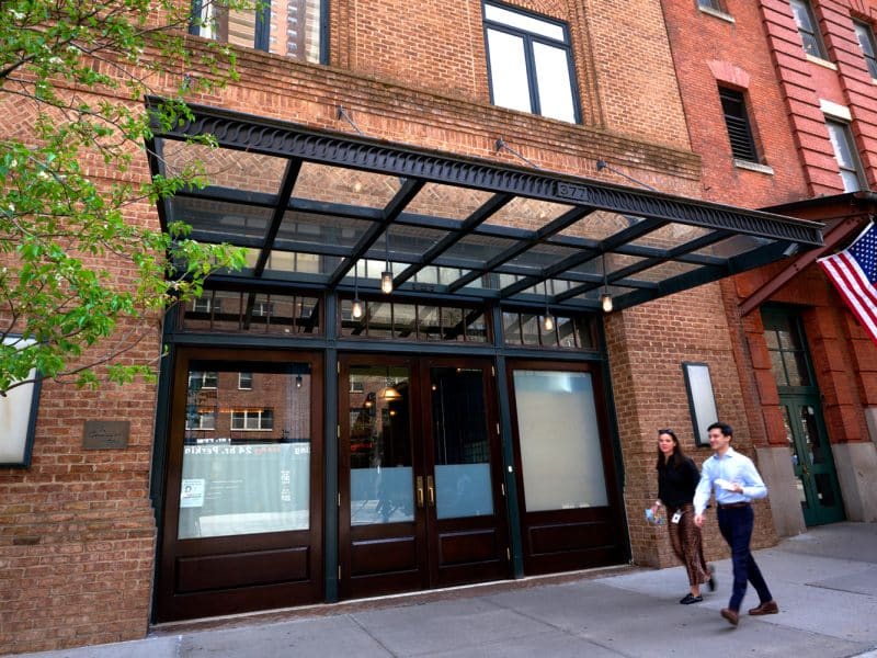 Entrance of The Greenwich Hotel in Tribeca, featuring a modern glass awning, brick facade, and two pedestrians walking by, with an American flag visible on the right.