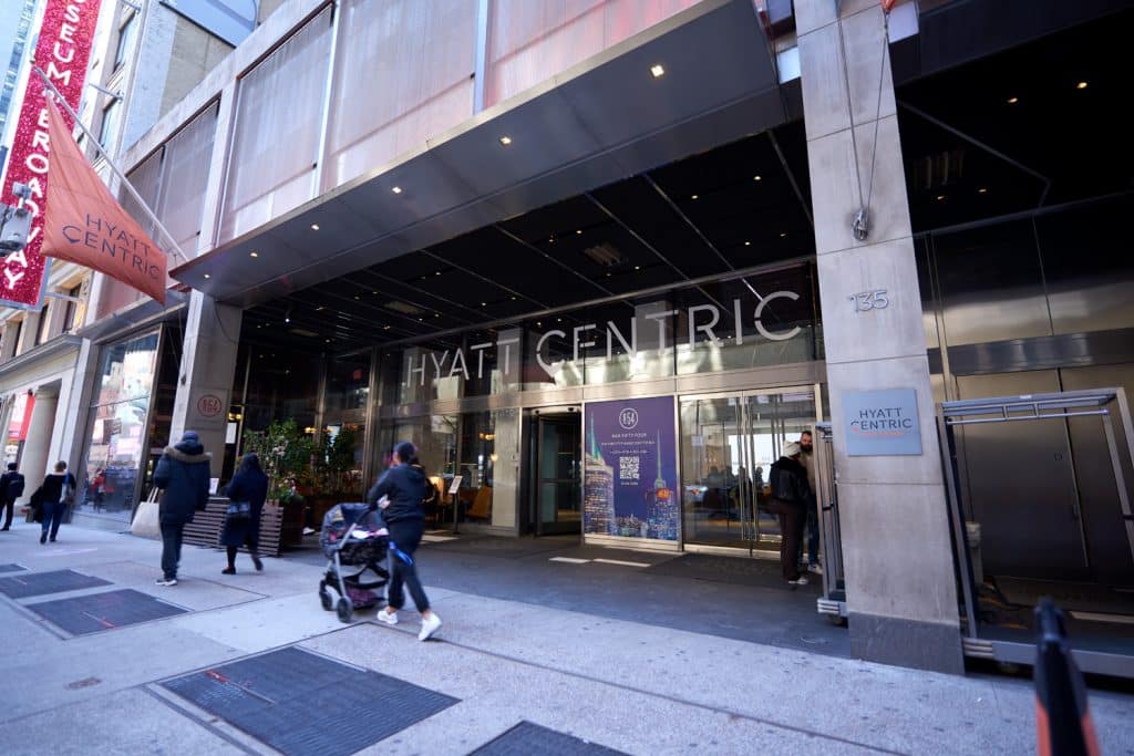 Hyatt Centric Times Square hotel entrance with modern architecture, large glass doors, and welcoming atmosphere, featuring guests arriving and a prominent brand logo.
