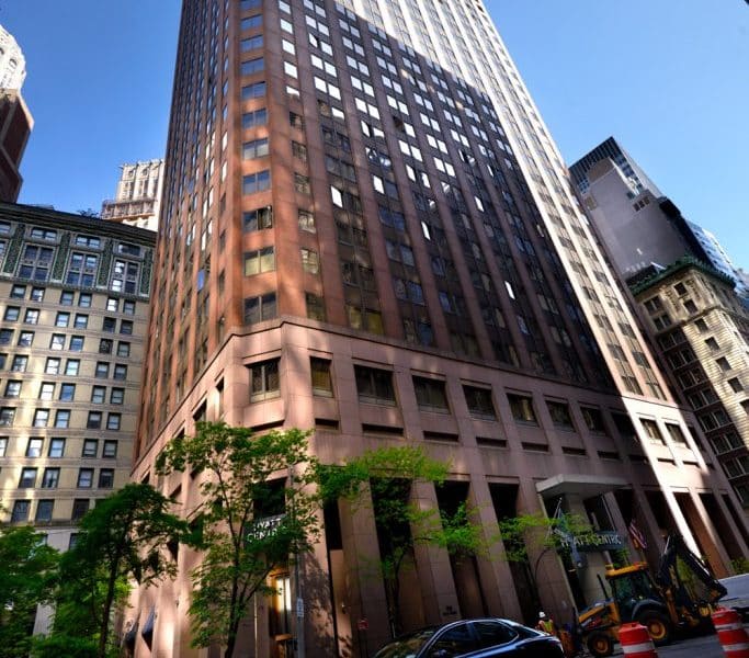 Hyatt Centric Wall Street hotel exterior in New York City's Financial District, showcasing a modern high-rise building with large windows, surrounded by greenery and nearby urban architecture.