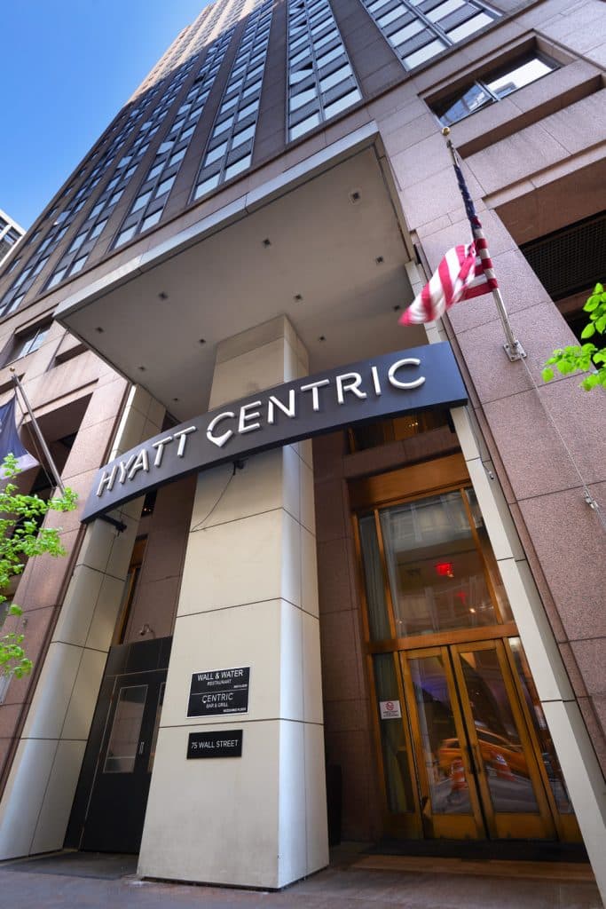 Hyatt Centric Wall Street hotel entrance with modern architecture, featuring the hotel name prominently displayed, American flag, and surrounding greenery.