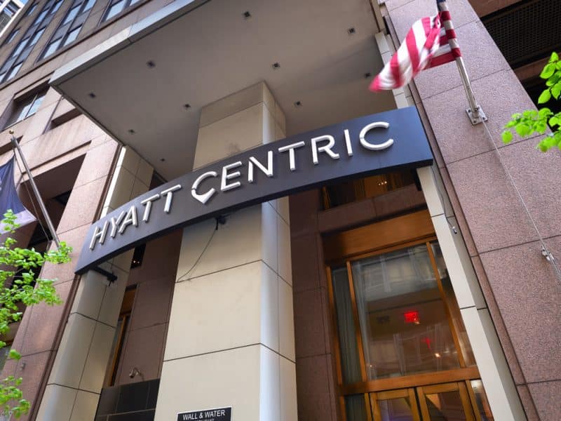 Hyatt Centric Wall Street hotel entrance with prominent signage, American flag, and surrounding greenery, reflecting a refined atmosphere in New York City's Financial District.