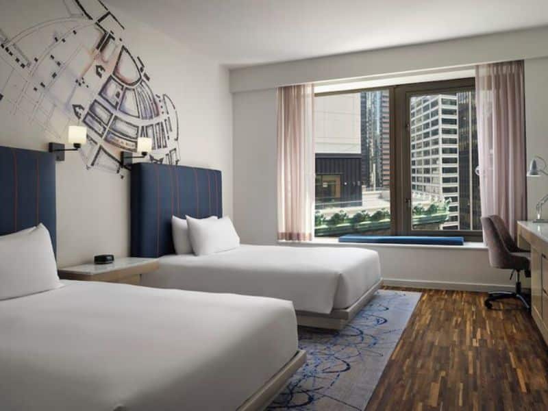 Modern hotel room at Hyatt Centric Wall Street featuring two beds, a workspace, and large window with city views, highlighting refined decor and comfort.