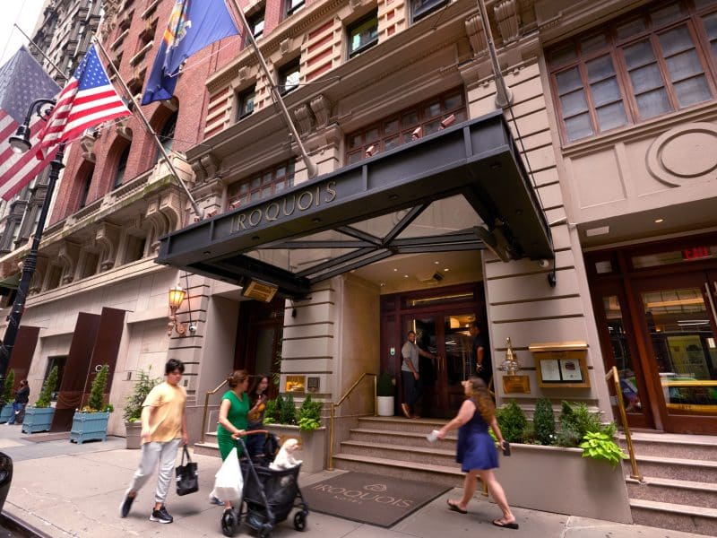 Iroquois Hotel entrance with flags, showcasing boutique luxury in Midtown Manhattan, featuring guests arriving and departing.