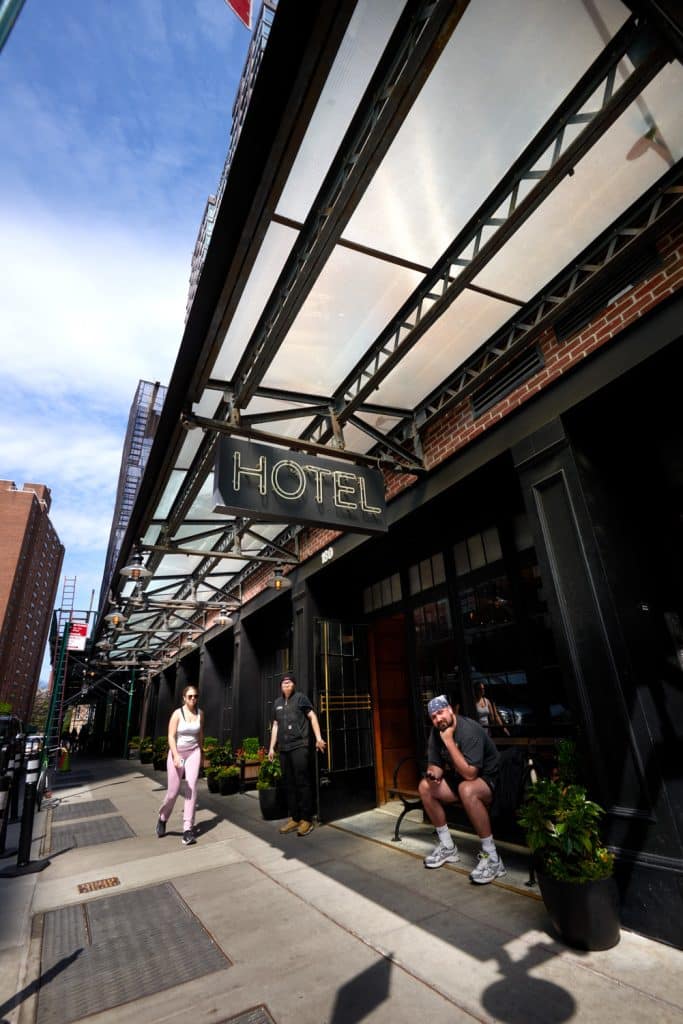 Boutique hotel entrance of The Ludlow Hotel in Manhattan's Lower East Side, featuring a stylish awning, "HOTEL" sign, and guests, including a woman in pink leggings and a staff member, surrounded by potted plants.