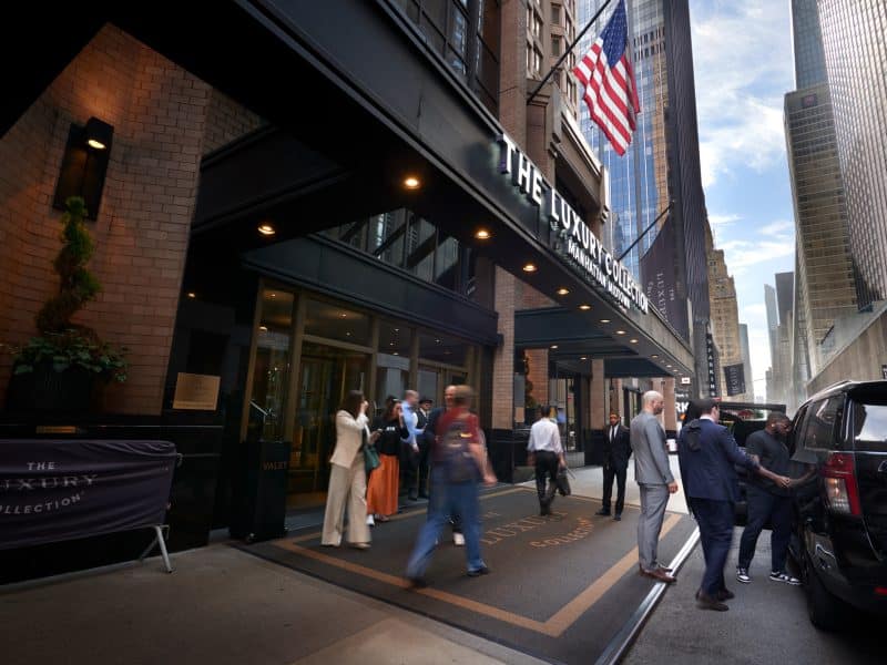 The Luxury Collection Hotel Manhattan Midtown entrance with guests arriving, valet service, and American flag, showcasing luxury accommodations in New York City.