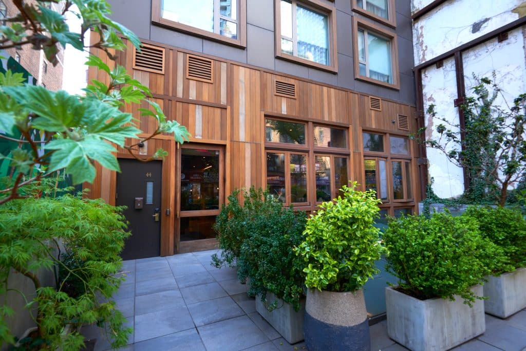 Entrance of Made Hotel featuring wooden paneling, large windows, and greenery, located in Manhattan's NoMad neighborhood.