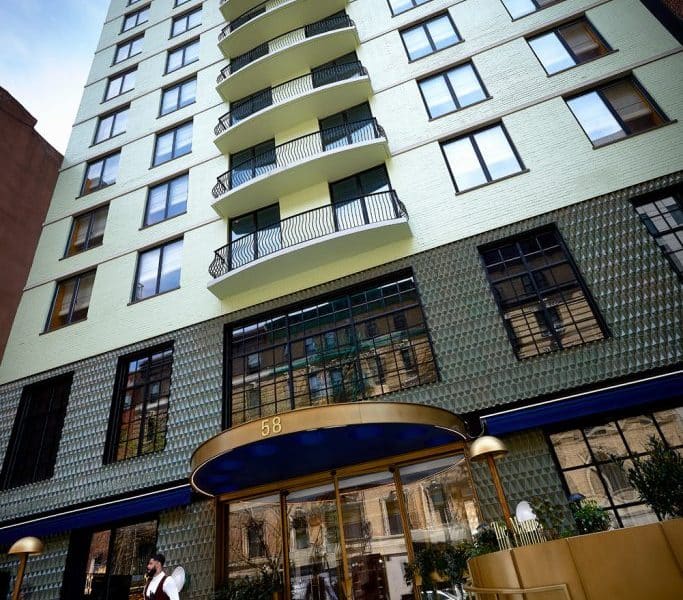 Chic boutique hotel exterior of The Manner Soho in Manhattan, featuring modern architecture, large windows, and a welcoming entrance with a gold canopy and potted plants.