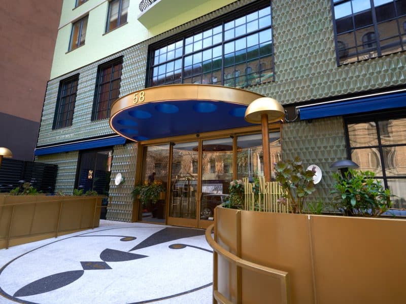The Manner Soho hotel entrance featuring modern design elements, gold awning, and lush greenery in Manhattan's SoHo neighborhood.