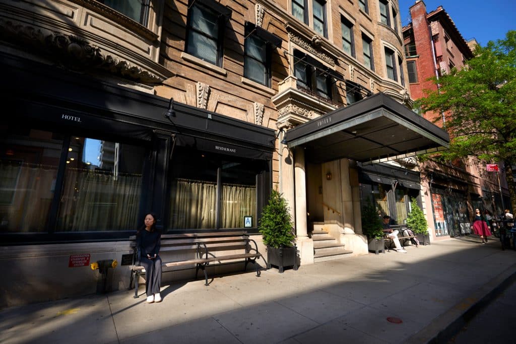 The Marlton Hotel entrance in Greenwich Village, featuring historic Beaux-Arts architecture, restaurant signage, and a woman sitting on a bench, reflecting the hotel's boutique charm and inviting atmosphere.