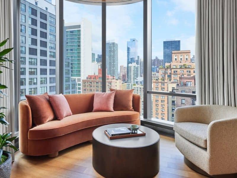 Luxury hotel room at Pendry Manhattan West, featuring a modern curved sofa with pink cushions, a round coffee table, and expansive windows showcasing the vibrant Manhattan skyline.