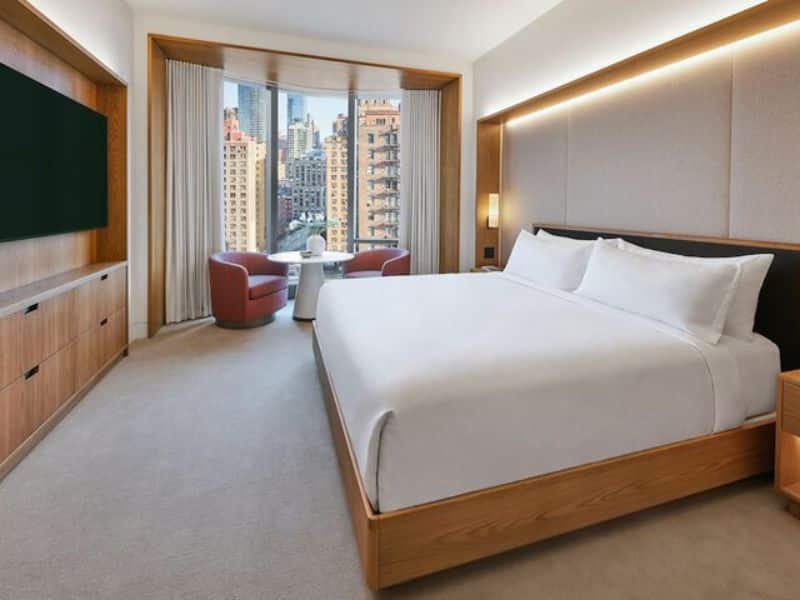 Luxury hotel room at Pendry Manhattan West featuring a king-sized bed, modern decor, and a view of the city skyline through large windows, with seating area including red chairs and a small table.