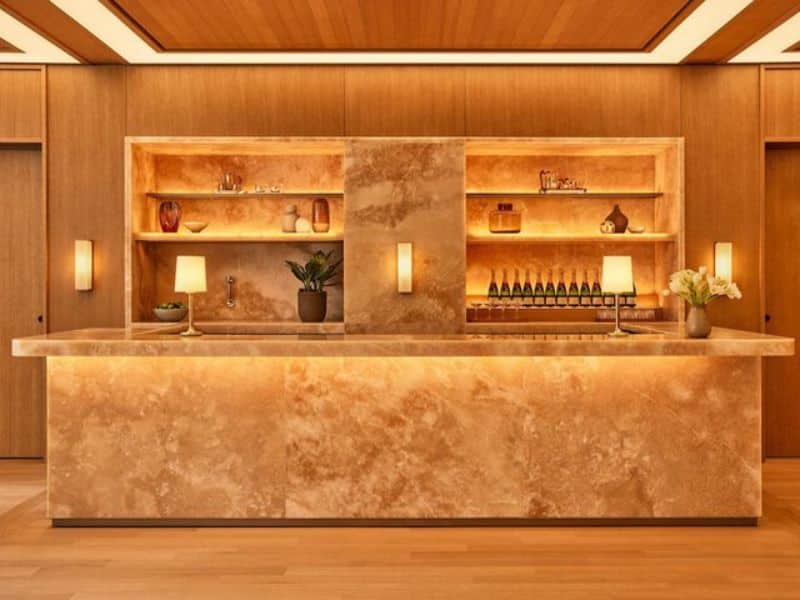 Luxury hotel reception area at Pendry Manhattan West, featuring a warm, contemporary design with a marble counter, illuminated shelves displaying decorative items and beverages, and elegant lighting.