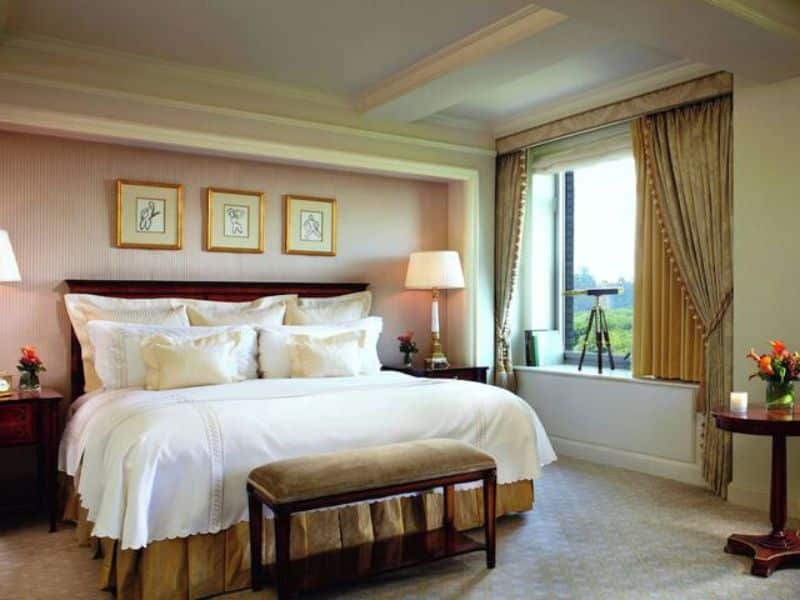 Luxurious hotel room at Ritz-Carlton Central Park featuring a king-size bed, elegant decor, and views of Central Park through large windows, complemented by a telescope and tasteful furnishings.