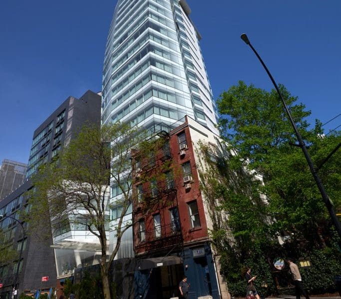 Modern boutique hotel exterior, The Standard – East Village, featuring a sleek glass tower alongside historic red brick building, surrounded by greenery and urban street scene.
