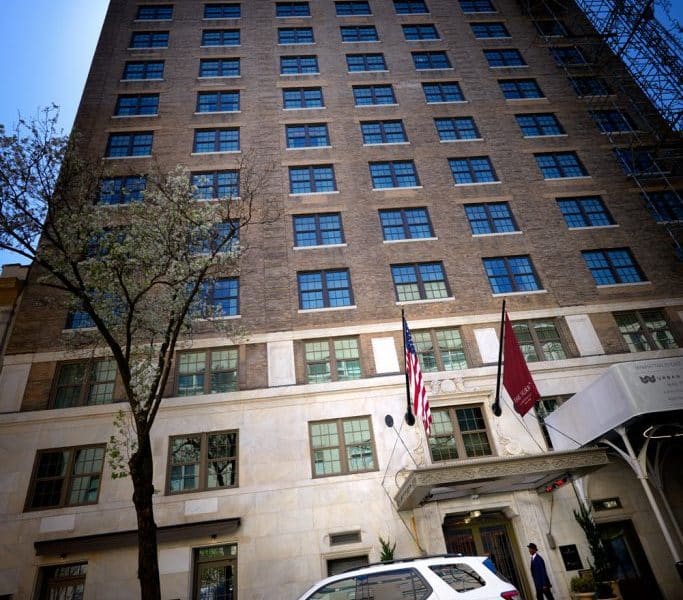 Exterior view of The Surrey, A Corinthia Hotel, showcasing the elegant brick facade, large windows, entrance canopy, American and hotel flags, and surrounding trees in Manhattan's Upper East Side.