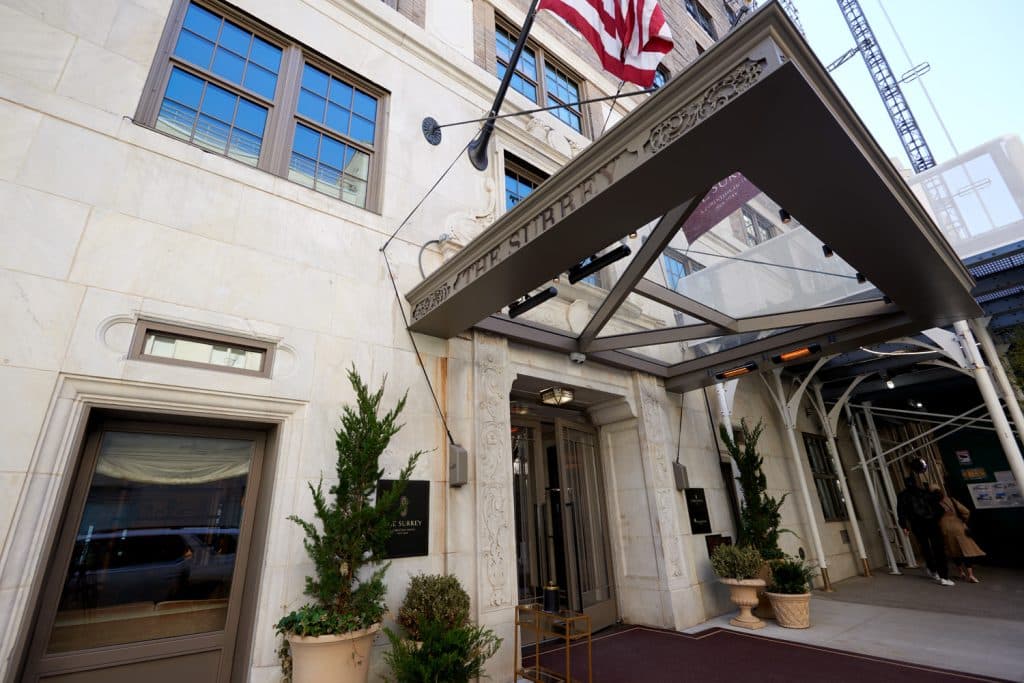 Entrance of The Surrey, A Corinthia Hotel, featuring elegant architecture, an awning, and potted greenery, with a flag and nearby pedestrians in Manhattan's Upper East Side.