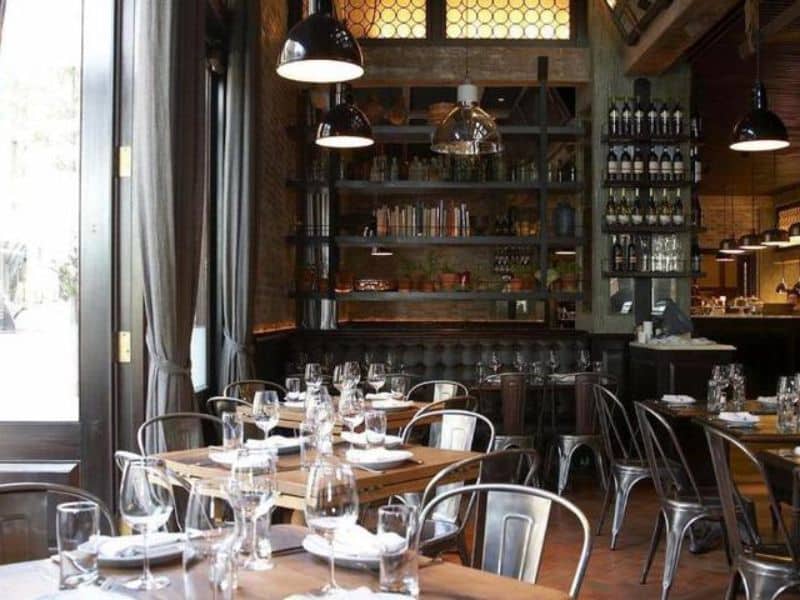Interior of The Greenwich Hotel restaurant featuring wooden tables set with glassware, industrial-style seating, and a stylish bar with shelves of wine and books.