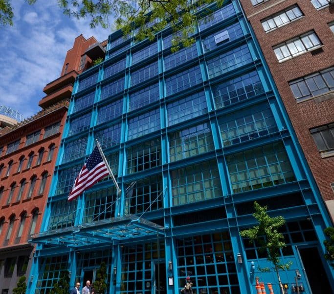 Warren Street Hotel exterior featuring modern blue glass façade, American flag, and entrance with greenery, located in Tribeca, New York City.