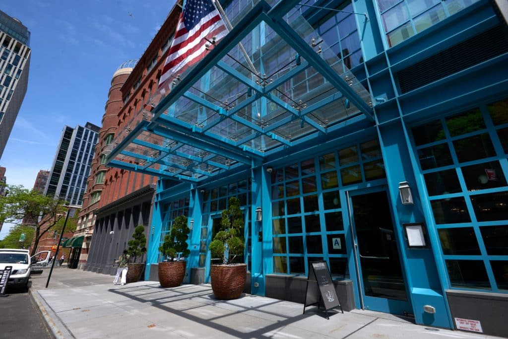 Warren Street Hotel entrance featuring modern glass architecture, vibrant blue exterior, and American flag, surrounded by urban Tribeca landscape.