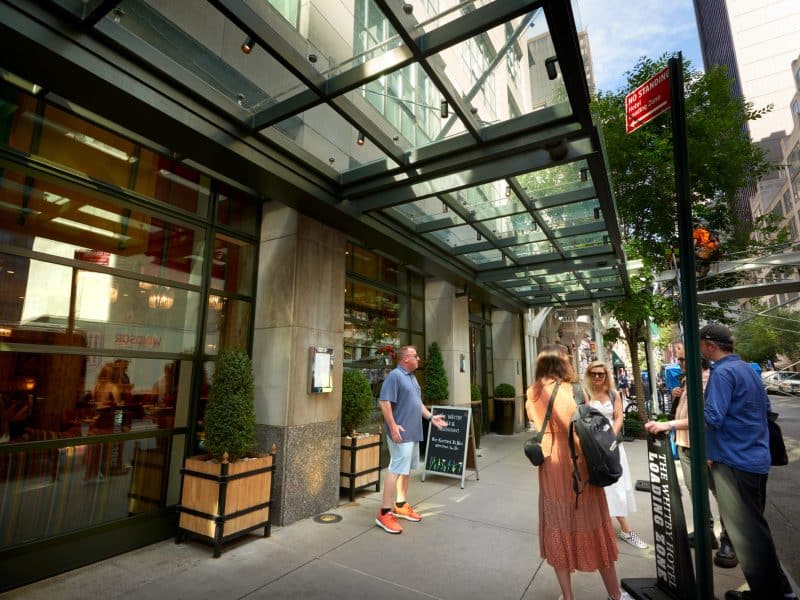 Entrance of The Whitby Hotel in Midtown Manhattan, featuring contemporary glass architecture, guests interacting, and a sidewalk menu display highlighting dining options.