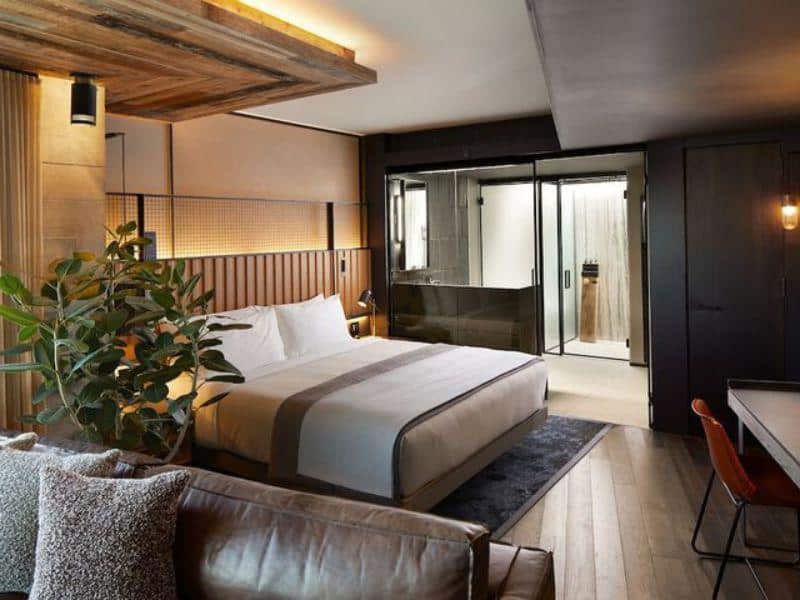 Luxurious hotel room at 1 Hotel Brooklyn Bridge featuring a king bed, modern decor, a cozy seating area, and a glass-enclosed bathroom, emphasizing eco-conscious design and comfort.