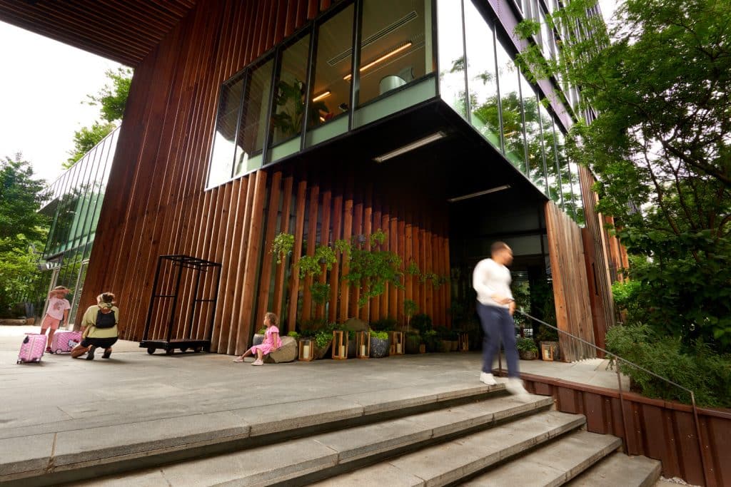 Family arriving at 1 Hotel Brooklyn Bridge, featuring modern eco-friendly architecture with wooden accents and greenery, children with luggage, and a man walking down the steps.