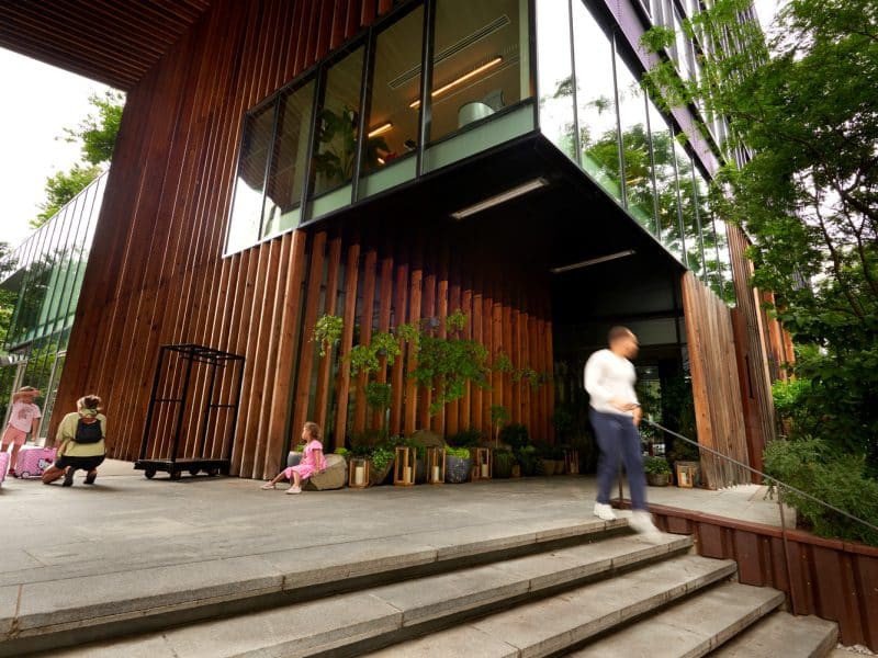 1 Hotel Brooklyn Bridge entrance with modern wooden architecture, lush greenery, and guests interacting, showcasing eco-friendly design and luxurious ambiance.