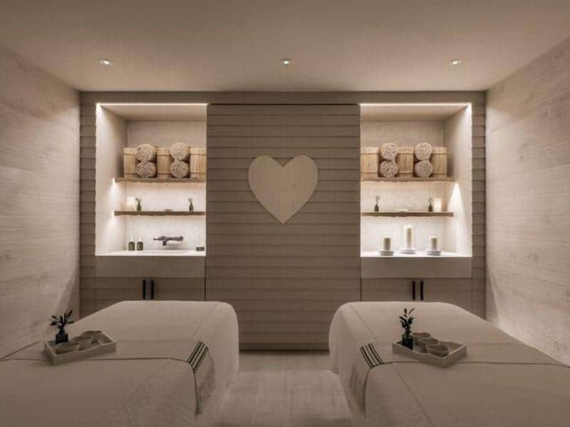 Spa treatment room at 1 Hotel Brooklyn Bridge featuring massage tables, wooden decor, heart-shaped wall art, and calming ambiance, highlighting eco-conscious luxury and wellness offerings.