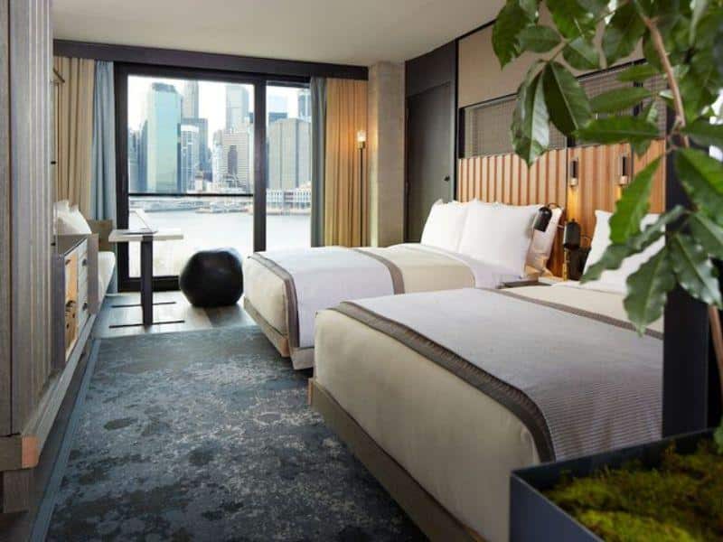Luxurious hotel room at 1 Hotel Brooklyn Bridge with two beds, eco-friendly design, large windows showcasing Manhattan skyline views, and modern decor elements.