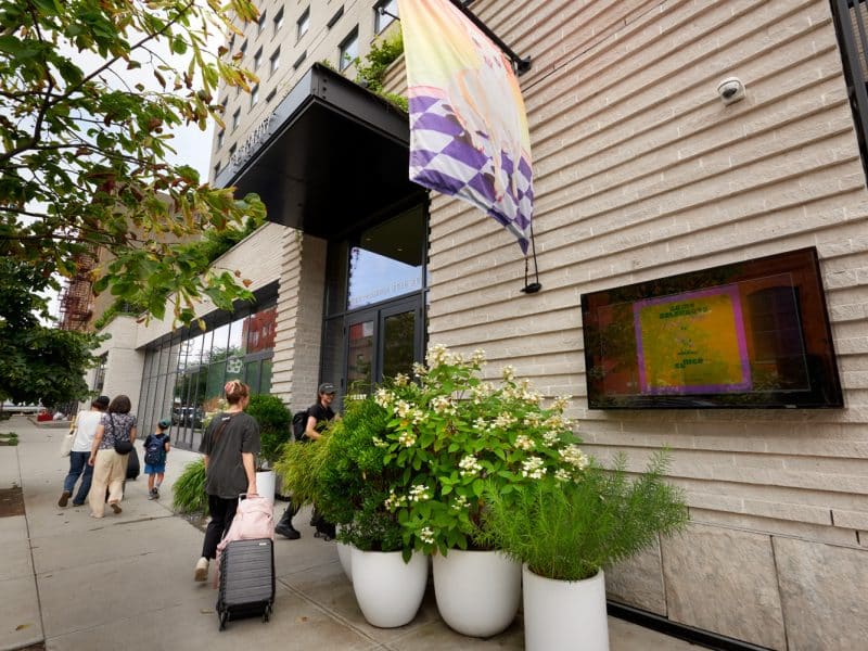 Penny Williamsburg hotel entrance with guests arriving, vibrant flag, modern architecture, and lush greenery in planters.