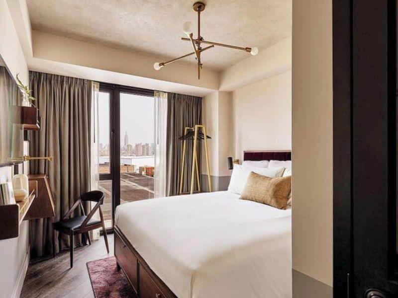 Stylish hotel room at The Hoxton, Williamsburg featuring modern design, luxury bed, and views of Manhattan skyline through large windows.