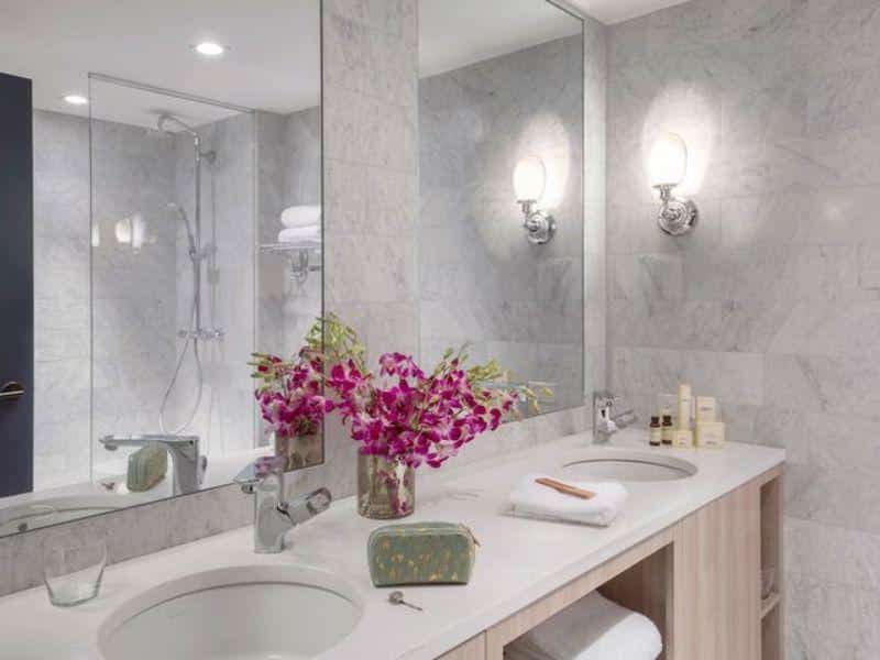 Modern hotel bathroom with marble walls, stylish fixtures, floral arrangement, toiletries, and ample lighting at The Tillary Hotel in Brooklyn.