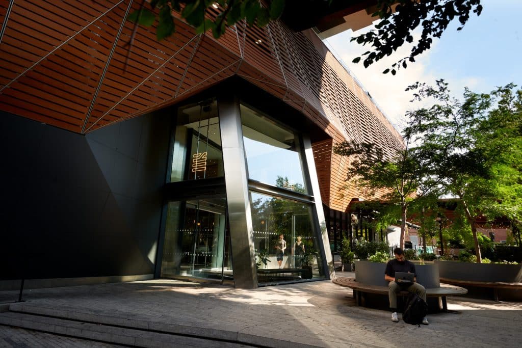 Modern architectural facade of The William Vale hotel in Williamsburg, Brooklyn, featuring a sleek entrance, wooden accents, and surrounding greenery, with a guest seated on a bench.