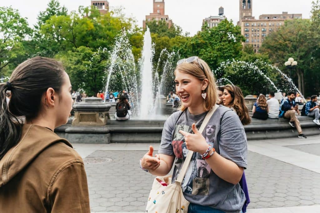 Two young women engaged in conversation near a fountain in Washington Square Park, with greenery and people in the background, reflecting a lively atmosphere of a guided tour exploring Taylor Swift hotspots in NYC.