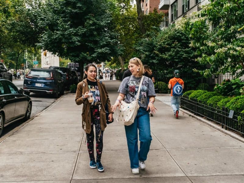 Two young women wearing Taylor Swift-themed apparel walking and chatting on a sidewalk in New York City, surrounded by trees and parked cars, while a person in an orange shirt walks in the background.