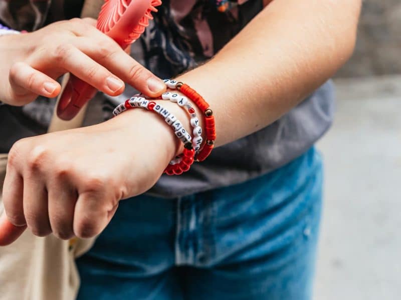 Person wearing colorful bracelets, including red and white beaded designs, pointing to wrist, highlighting Taylor Swift fandom during NYC tour.