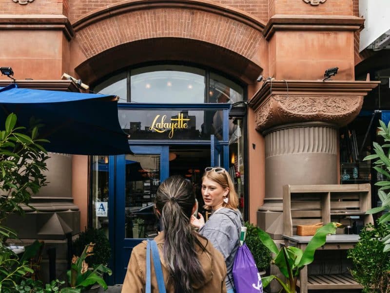 Women in front of Lafayette Grand Caf&eacute; & Bakery, a notable stop on the Taylor Swift hotspots tour in NYC, showcasing the entrance and surrounding greenery.