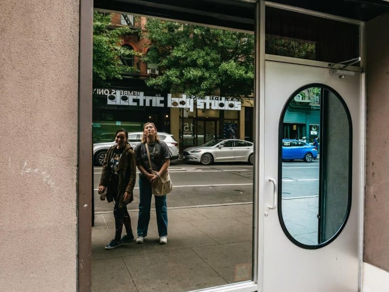Two women posing in front of a reflective storefront window, capturing their visit to Taylor Swift hotspots in NYC, with urban scenery and "Electric Lady Studios" visible in the background.