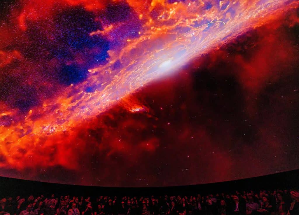 Audience watching a vibrant planetarium show featuring a colorful cosmic scene, showcasing swirling galaxies and nebulae, at the American Museum of Natural History.