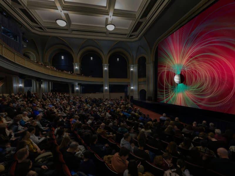 Crowd gathered in the Rose Center for Earth and Space at the American Museum of Natural History, with a vibrant projection on the screen depicting celestial visuals.