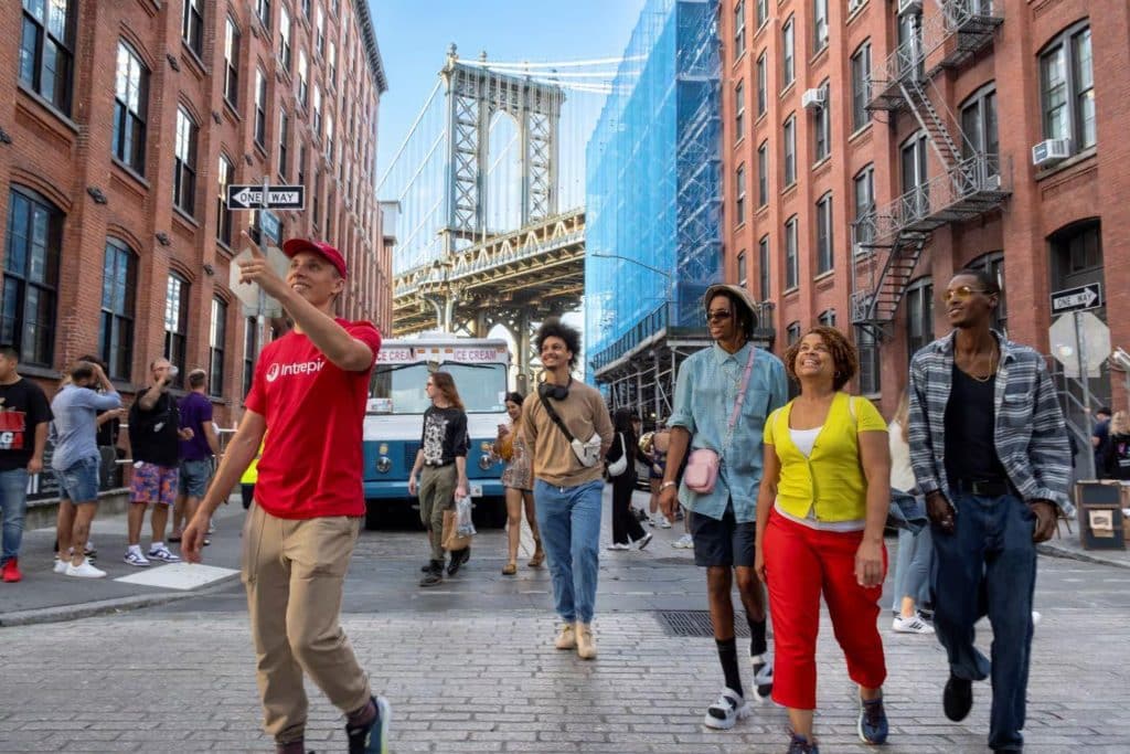 Group of diverse people enjoying a food tour in DUMBO, Brooklyn, with the Manhattan Bridge in the background, capturing the vibrant culinary scene and local culture.