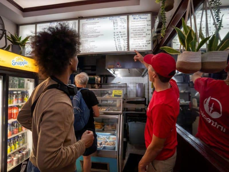 Person pointing at a menu in a Brooklyn eatery, showcasing diverse food options, with patrons engaged in a culinary experience during the Brooklyn Heights and DUMBO Neighborhood Food Tour.