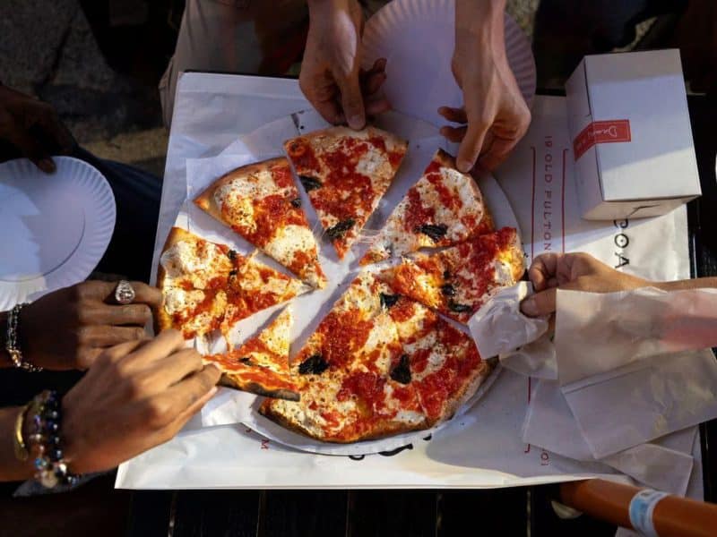 Hands reaching for slices of pizza on a paper plate, showcasing a communal dining experience during the Brooklyn Heights and DUMBO Neighborhood Food Tour.