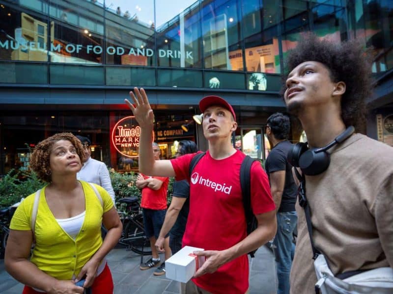 Group of diverse food tour participants listening to a guide in a red shirt, with the Museum of Food and Drink and Time Out Market in the background, highlighting Brooklyn's culinary scene.