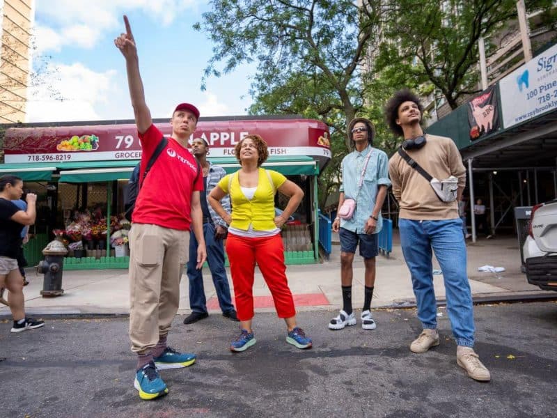 Group of diverse individuals exploring Brooklyn Heights, gesturing and looking up, with a local grocery store in the background, showcasing the vibrant atmosphere of the neighborhood food tour.