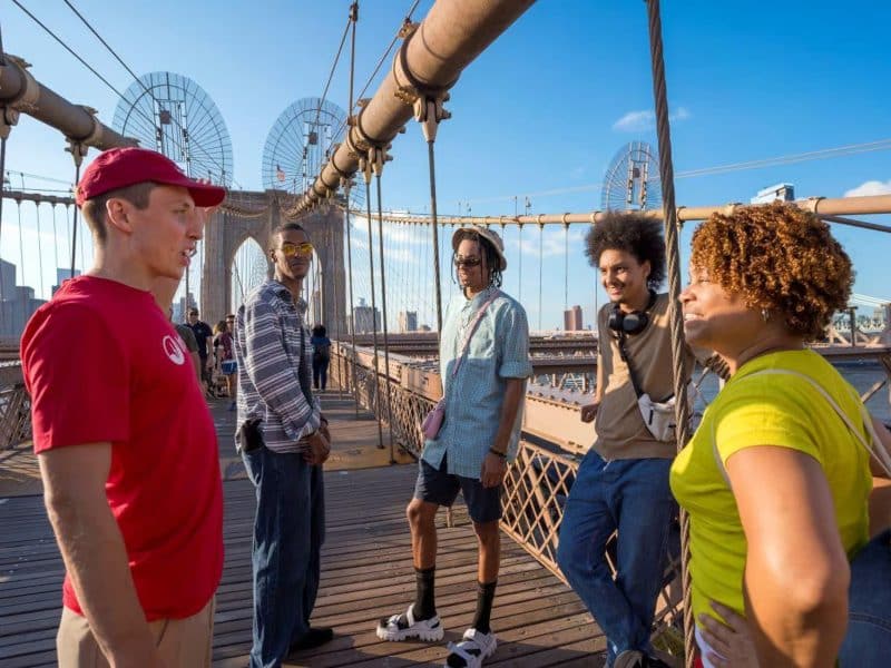 Group of diverse individuals engaging in conversation on the Brooklyn Bridge during a food tour, with iconic bridge architecture and Manhattan skyline in the background.